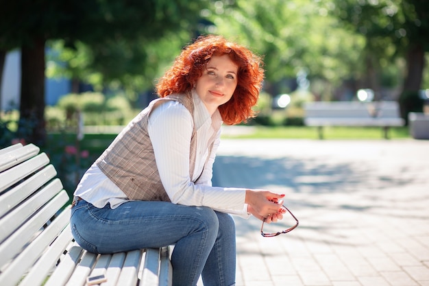 Schöne rothaarige Frau sitzt auf einer Bank im Park an einem sonnigen Sommertag und genießt das Leben