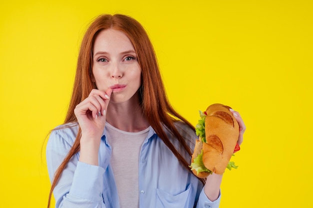 Schöne rothaarige Ingwerfrau echte Marmelade, die Finger leckt, die ein riesiges Sandwich Titanic im gelben Hintergrund des Studios halten.