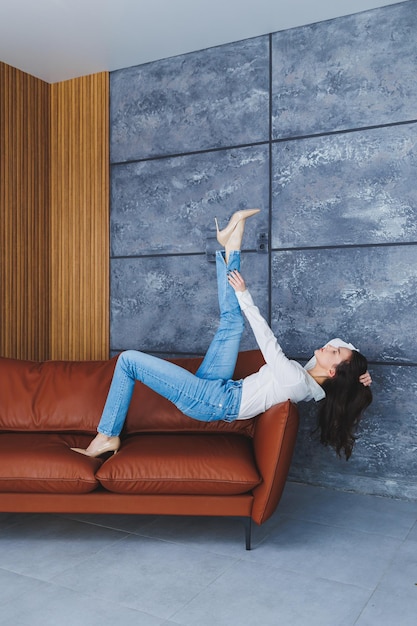 Schöne schlanke Frauenbeine in klassischen beigen Schuhen mit dünnen Absätzen und Jeans Stilvolles Mädchen in Absätzen sitzt auf einem braunen Ledersofa