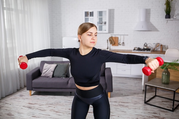 Schöne schlanke junge Frau treibt zu Hause in einem Top und Leggings Sport. Fitness zu Hause für einen schönen Körper. Übungen mit Hanteln für den weiblichen Körper. Gesunder Lebensstil.