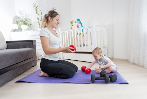 Schöne schlanke mutter mit ihrem kleinen sohn, der zu hause auf einer fitnessmatte sitzt und mit hanteln trainiert