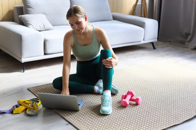 Schöne schlanke sportliche Frau in Sportkleidung sitzt mit Hanteln auf dem Boden und benutzt einen Laptop zu Hause im Wohnzimmer. Gesunder Lebensstil. Bleiben Sie zu Hause Aktivitäten.