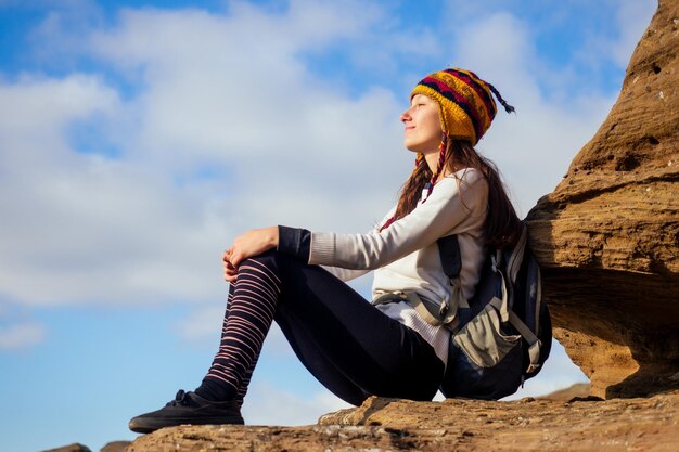 Schöne schlanke und sportliche junge Touristenfrau in einem lustigen Hut aus nepalesischem Wollyak sitzt und ruht sich beim Klettern großer Felsen auf Felsbrocken im Canyon-Steine-Himmel aus