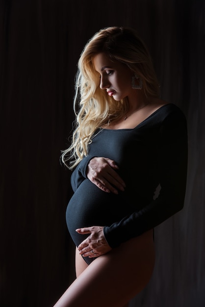 Schöne schwangere blonde Frau steht und umarmt ihren Bauch an einer schwarzen Wand.
