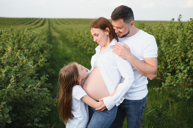 Schöne schwangere brünette Mutter und Vater und Tochter in weißen Kleidern sehen sich in der Natur an