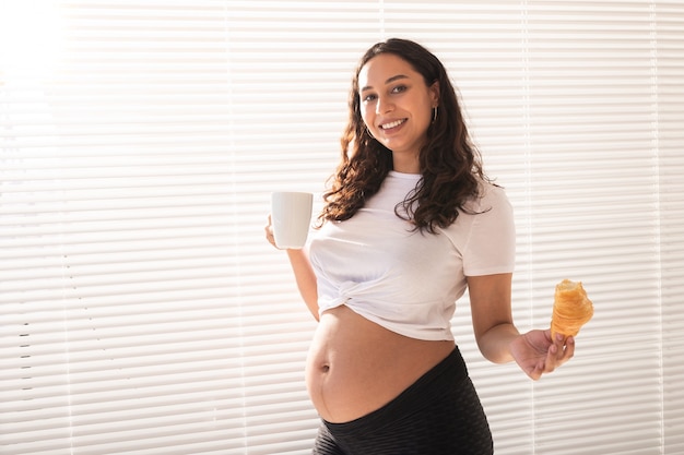 Schöne schwangere Frau, die Croissant und Tasse Kaffee in ihren Händen während des Morgenfrühstücks hält. Konzept der guten Gesundheit und der positiven Einstellung beim Erwarten des Babys