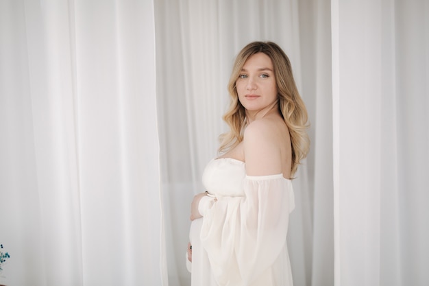 Schöne schwangere Frau im eleganten weißen Kleid posiert für den Fotografen im Studiohintergrund von Weiß