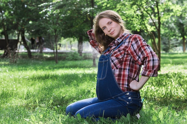 Schöne schwangere Frau in Denim-Overalls, die auf dem Gras im Park sitzt