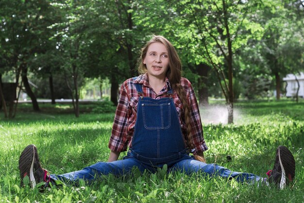 Schöne schwangere Frau in Denim-Overalls, die auf dem Gras im Park sitzt