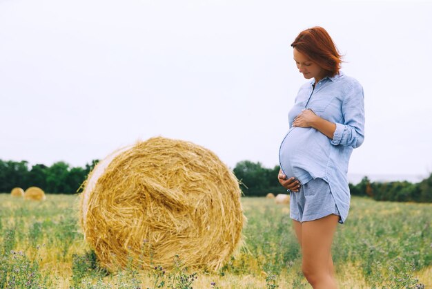 Schöne schwangere Frau in der Natur im Freien Frau hält die Hände auf Bauch auf Hintergrund des Weizenfeldes mit Heuhaufen am Sommertag Foto der Schwangerschaft Mutterschaftserwartung Mutter wartet auf ein Baby