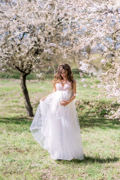 Schöne schwangere Frau in einem langen hellen Kleid geht in einem blühenden Frühlingsgarten.