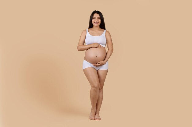 Schöne schwangere Frau in Unterwäsche, die den Bauch zärtlich umarmt und in die Kamera lächelt