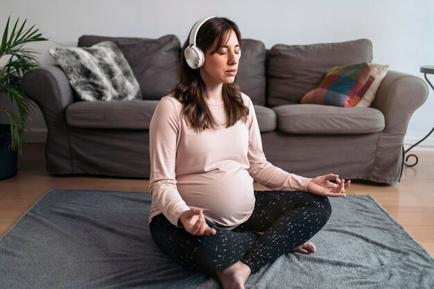 Schöne schwangere Frau macht zu Hause Yoga, hört Musik und meditiert