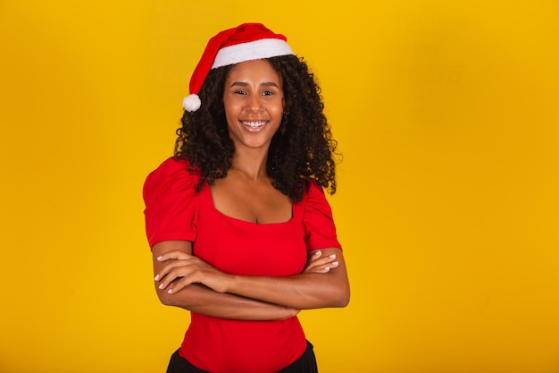 Schöne schwarze Afrofrau mit lockigem Haar auf gelbem Hintergrund in Weihnachtsmütze mit verschränkten Armen und freiem Platz für Text, der in die Kamera lächelt