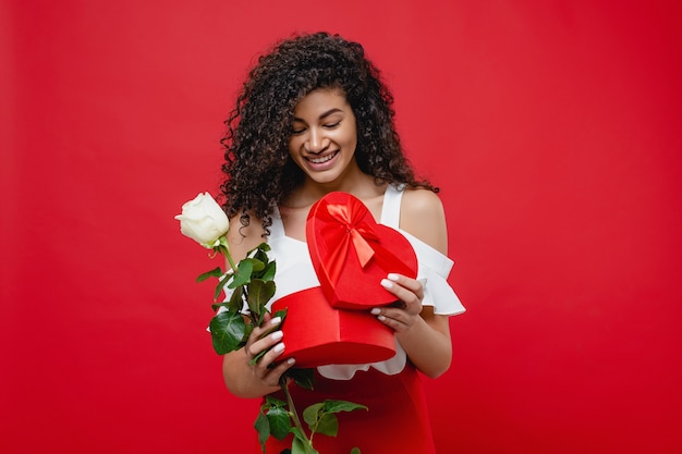 Schöne schwarze Frau, die Herzformgeschenk und Weißrose lokalisiert auf Rot lächelt und hält