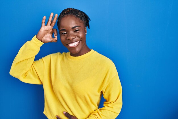Schöne schwarze Frau, die über blauem Hintergrund steht und positiv lächelt und mit Hand und Fingern ein erfolgreiches Ausdruckszeichen macht