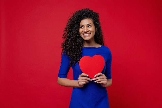 Schöne schwarze Frau im blauen Kleid mit Herzen formte Valentinsgrußkarte auf roter Wand