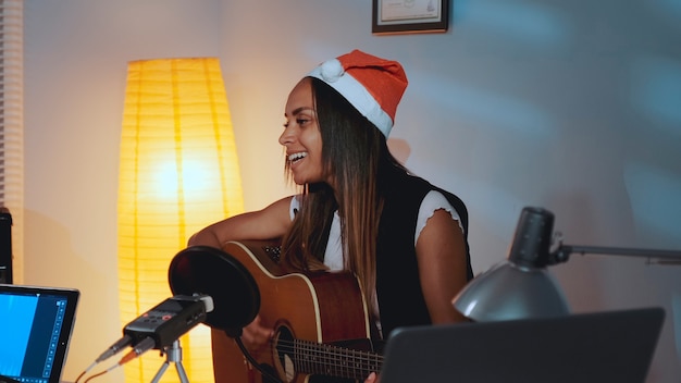 Schöne schwarze Frau in Weihnachtsmütze singt und spielt Gitarre an Heiligabend im gemütlichen Heimstudio i...