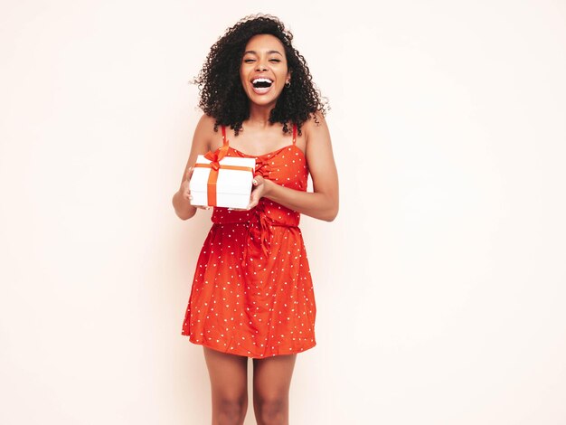 Schöne schwarze Frau mit Afro-Locken-Frisur Lächelndes Modell in rotem Sommerkleid gekleidet Sexy sorglose Frau posiert in der Nähe einer weißen Wand im Studio Gebräunt und fröhlich Holding-Geschenkbox