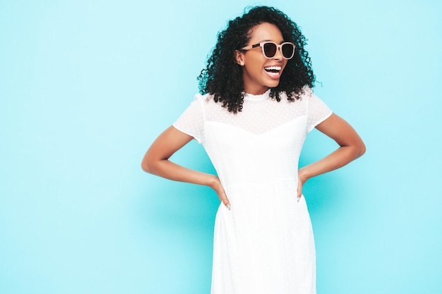 Schöne schwarze Frau mit Afro-Locken-Frisur Lächelndes Modell in weißem Sommerkleid gekleidet Sexy sorglose Frau posiert in der Nähe der blauen Wand im Studio Gebräunt und fröhlich In Sonnenbrille Isoliert