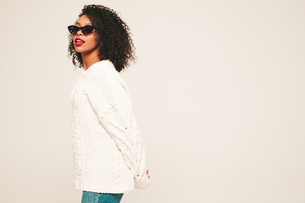 Schöne schwarze Frau mit Afro-Locken-Frisur und roten Lippen. Lächelndes Modell in trendiger Jeanskleidung und Winterpullover.