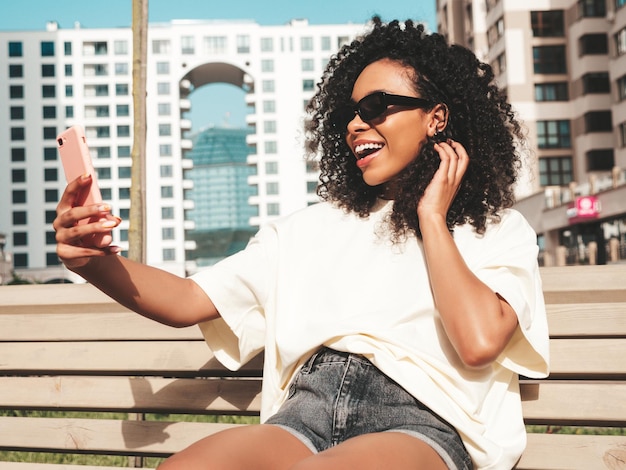 Schöne schwarze Frau mit Afro-Locken-FrisurLächelndes Hipster-Modell in weißem T-Shirt Sexy sorglose Frau, die mit Sonnenbrille auf der Straße posiert Fröhlich und glücklichSelfie-Foto machen