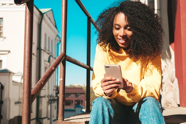 Schöne schwarze Frau mit Afro-Locken-FrisurLächelndes Modell in gelbem Hoodie Sexy sorglose Frau, die auf dem Straßenhintergrund posiert Blick auf den Smartphone-Bildschirm mit Apps