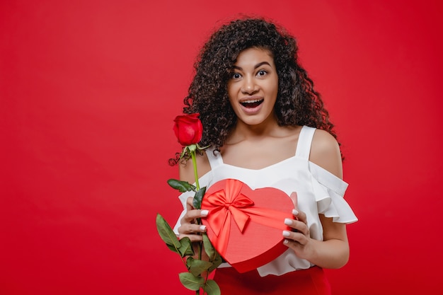 Schöne schwarze Frau mit Rose und Herz formte die Geschenkbox, die über Rot lokalisiert wurde