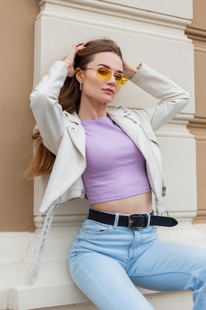 Schöne sexy Model-Frau in modischem, hellem Outfit mit weißer Lederjacke und Vintage-Blue-Jeans mit gelber Sonnenbrille sitzt und posiert in der Nähe der Wand neben dem Gebäude