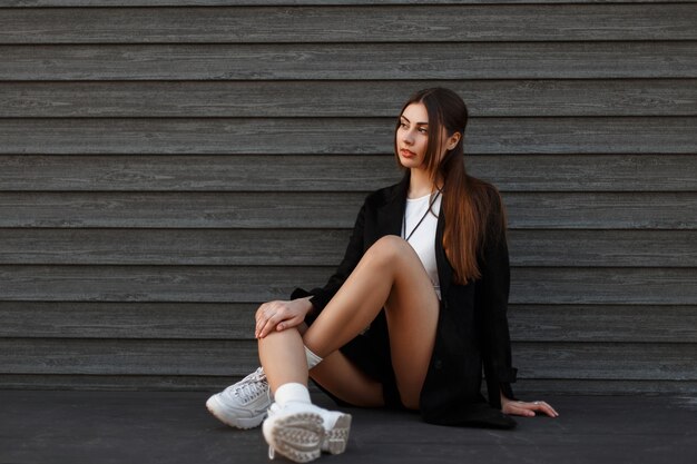 Schöne sexy Modellfrau im schwarzen Mantel mit modischen Schuhen, die nahe einer Holzwand sitzen