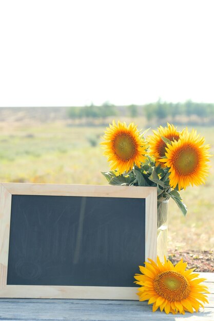 Foto schöne sonnenblumen in vase und leerem rahmen auf holztisch auf feldhintergrund