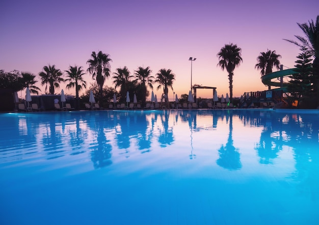 Schöne Spiegelung im Swimmingpool bei farbenprächtigem Sonnenuntergang Lila Himmel spiegelt sich im Wasser wider Palmen Sonnenliegen Sonnenschirme in der Nacht im Sommer Luxusresort Landschaft mit leerem Pool in der Dämmerung