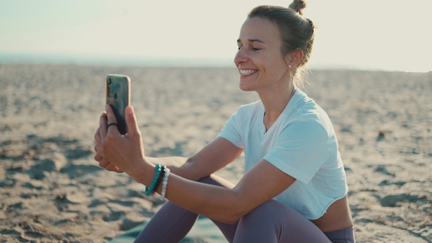 Foto schöne sportliche frau, die im video-chat über das smartphone spricht und sich nach dem yoga am strand ausruht junge frau, die ein mobiltelefon für die fernkommunikation verwendet