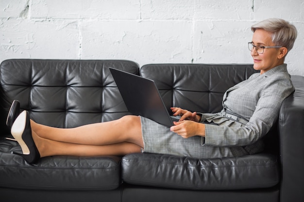 schöne stilvolle erwachsene Frau im Anzug, die mit Laptop auf schwarzem Sofa arbeitet