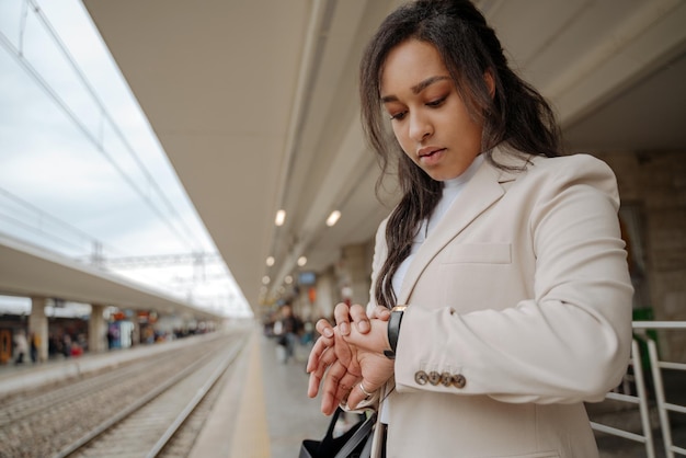 Schöne stilvolle Frau mit gemischter Rasse, die am Bahnhof steht und auf die Uhr schaut