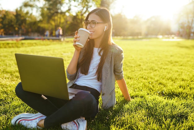 Schöne stilvolle Frau sitzt auf grünem Gras mit Laptop und Telefon in den Händen Lifestyle-Konzept