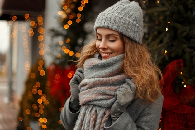 Schöne stilvolle junge Frau, die in gestrickten modischen Kleidern mit einer Mütze und einem Schal an Feiertagen nahe den Lichtern und einem Weihnachtsbaum an einem verschneiten Tag lächelt