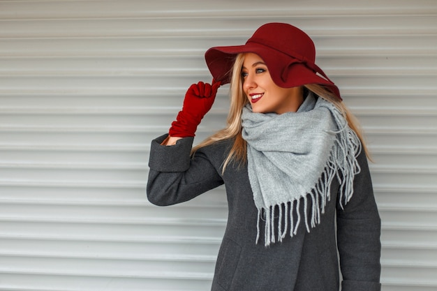 Foto schöne stilvolle junge frau in einem trendigen mantel mit einem grauen schal und einem eleganten hut, der nahe einer holzwand aufwirft