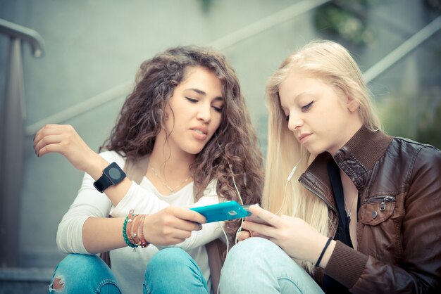 Schöne stilvolle junge Frauen der Blondine und des Brunette, die intelligentes Telefon verwenden