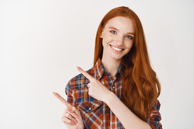 Schöne Studentin mit blasser Haut und langem natürlichen Ingwerhaar, die mit dem Finger nach links auf das Logo zeigt, das über weißem Hintergrund steht