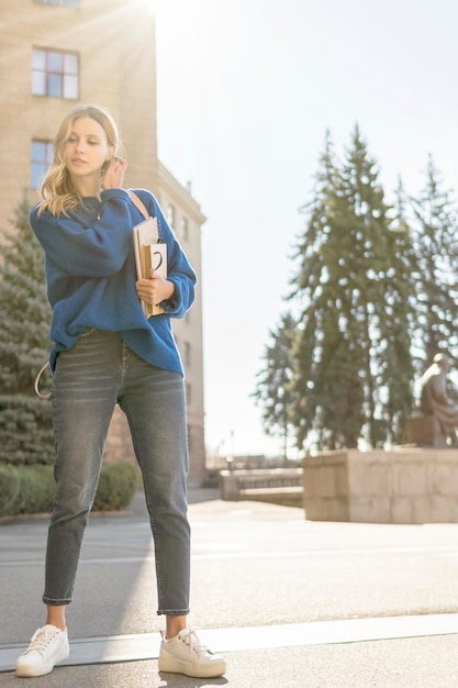 Schöne Studentin mit Notizbüchern in der Hand glättet an einem sonnigen Tag ihr Haar vor dem Hintergrund des Universitätsgebäudes