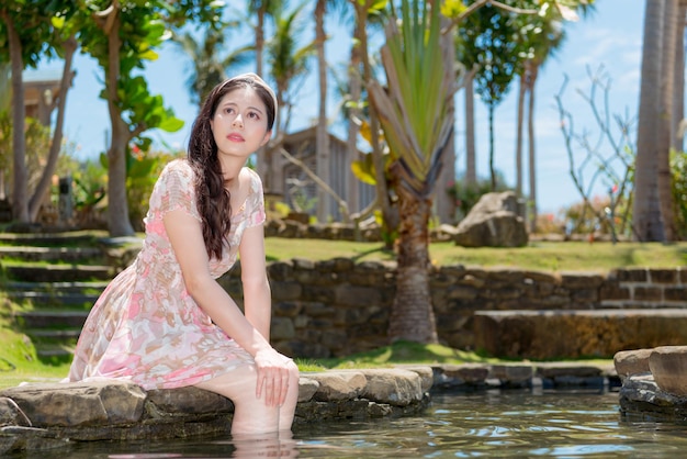 schöne süße dame, die sommerkleid trägt, das auf der teichseite sitzt, lässt beine im wasser entspannen und in der luft träumend denken reiseplanung.