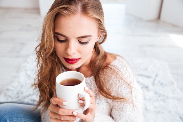 Schöne süße Frau mit langen Haaren und rotem Lippenstift, die Teetasse drinnen hält