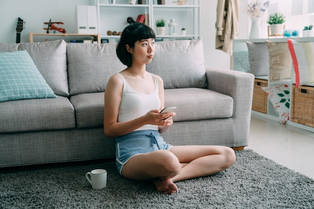 Schöne süße junge asiatische Chinesin mit dunklen kurzen Haaren, die Cappuccino genießt und mit dem Handy auf der Komfortetage im Wohnzimmer im Internet surft. Frau, die Seite schaut, genießt soziale Medien.