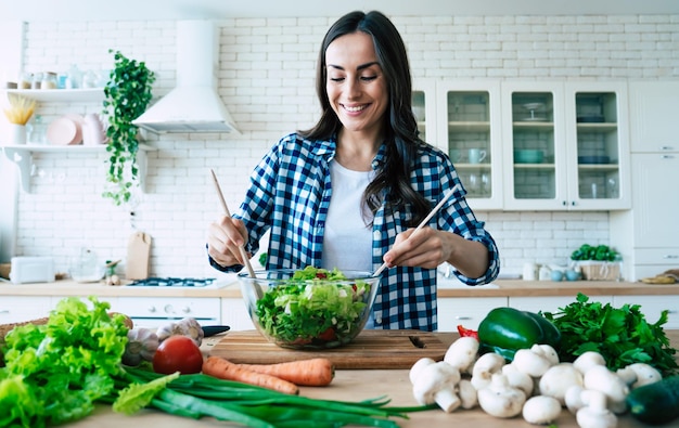 Schöne süße junge lächelnde Frau in der Küche bereitet einen veganen Salat in Freizeitkleidung zu