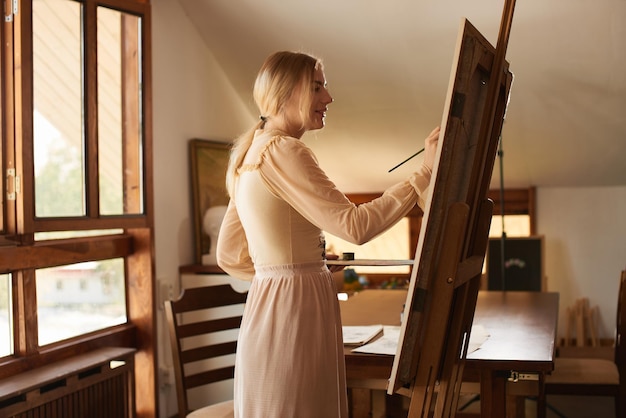 Schöne talentierte junge Künstlerin malt ein Bild mit Pinsel und Öl auf einer Staffelei mit Palette in der Hand in einem stilvollen kreativen Kunstatelier