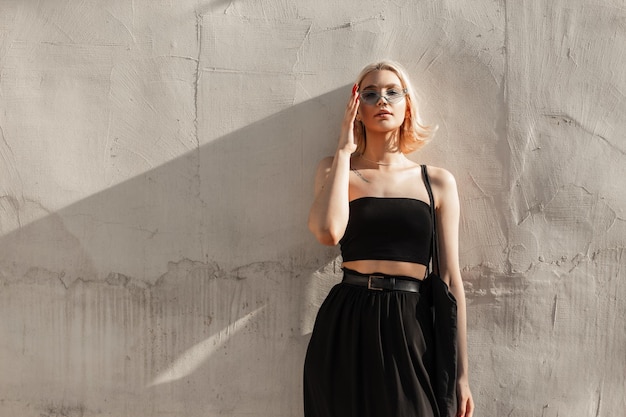 Schöne Teenager blonde Frau mit Sonnenbrille in Mode schwarzes Top und Rock steht in der Nähe einer Wand im Sonnenlicht