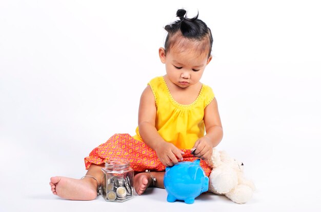 Schöne thailändische Baby-Mädchen sitzt und spielt Geldmünzen mit dem Sparschwein für Finanzideen-Konzept