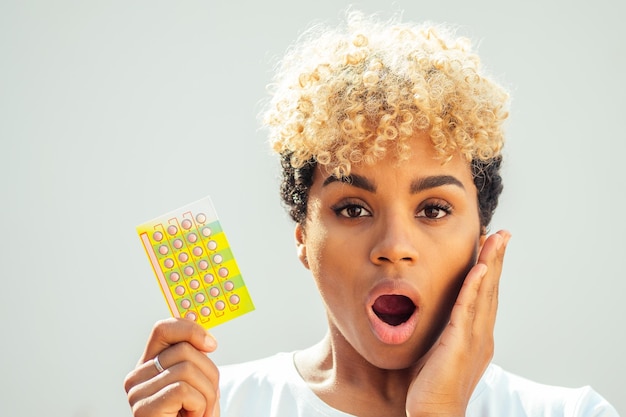 Schöne überraschende afroamerikanische Frau mit Antibabypille und offenem Mund, weil sie vergessen hat, die Pillen rechtzeitig einzunehmen Zahnschmerzkonzept