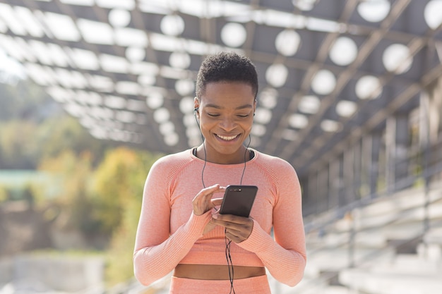 Schöne und erfolgreiche Sportlerin, die lächelt, hört während der morgendlichen Fitness und beim Joggen in der Nähe des Stadions ein Hörbuch vom Handy, afroamerikanische Frau mit Handy